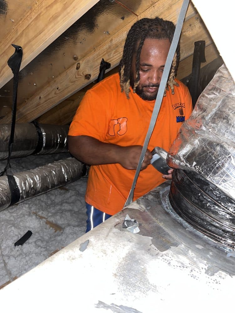 Focused man in an orange shirt repairs flexible HVAC ductwork in a cramped, insulated attic.
